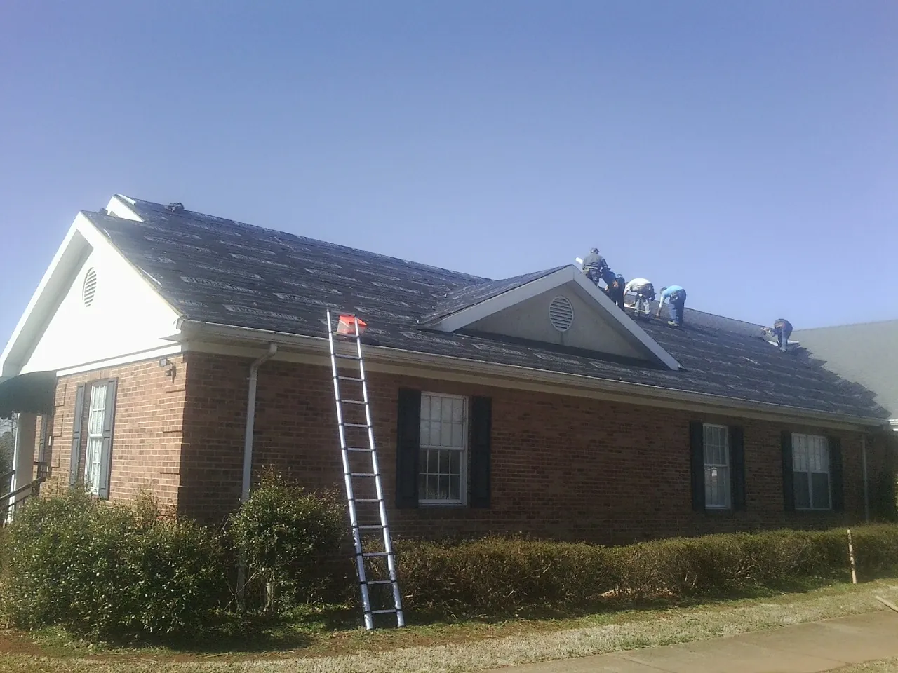 Crew tearing shingles off a brick ranch roof with a ladder set — Emergency Roof Repair work by our crew in Clute